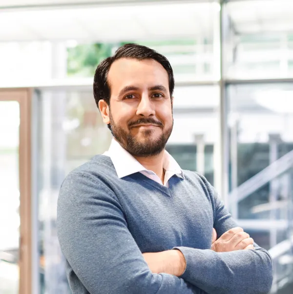 Head and body shot of Ammo. He is in a white shirt and smart blue jumper. He has a slight smile, arms folded, body angled to the right and he is looking directly at the camera.