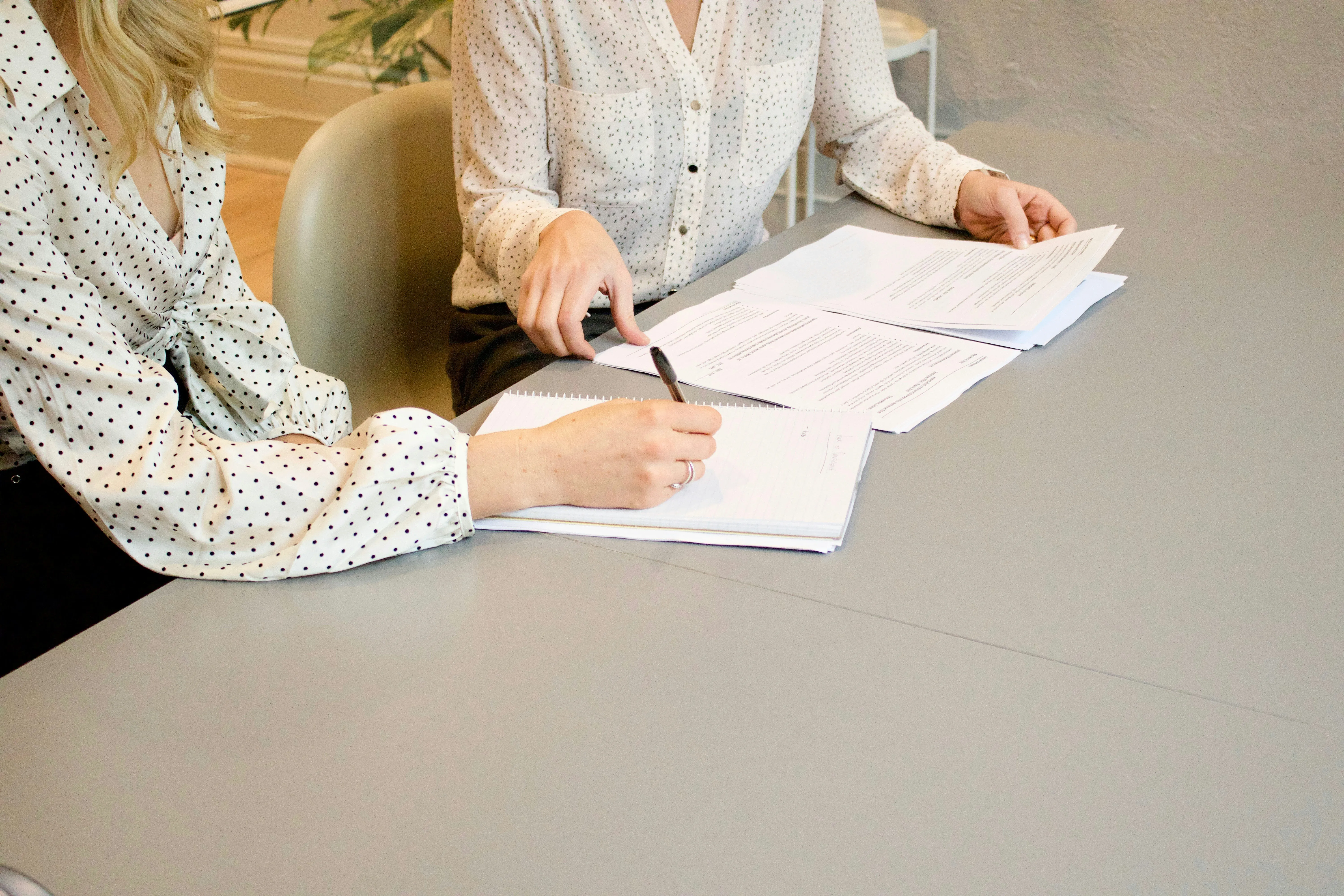 Two women sitting at a table. One is holding a pen above some paperwork ready to sign. The other is helping her by showing her where to sign and has further piles of paperwork in front.
