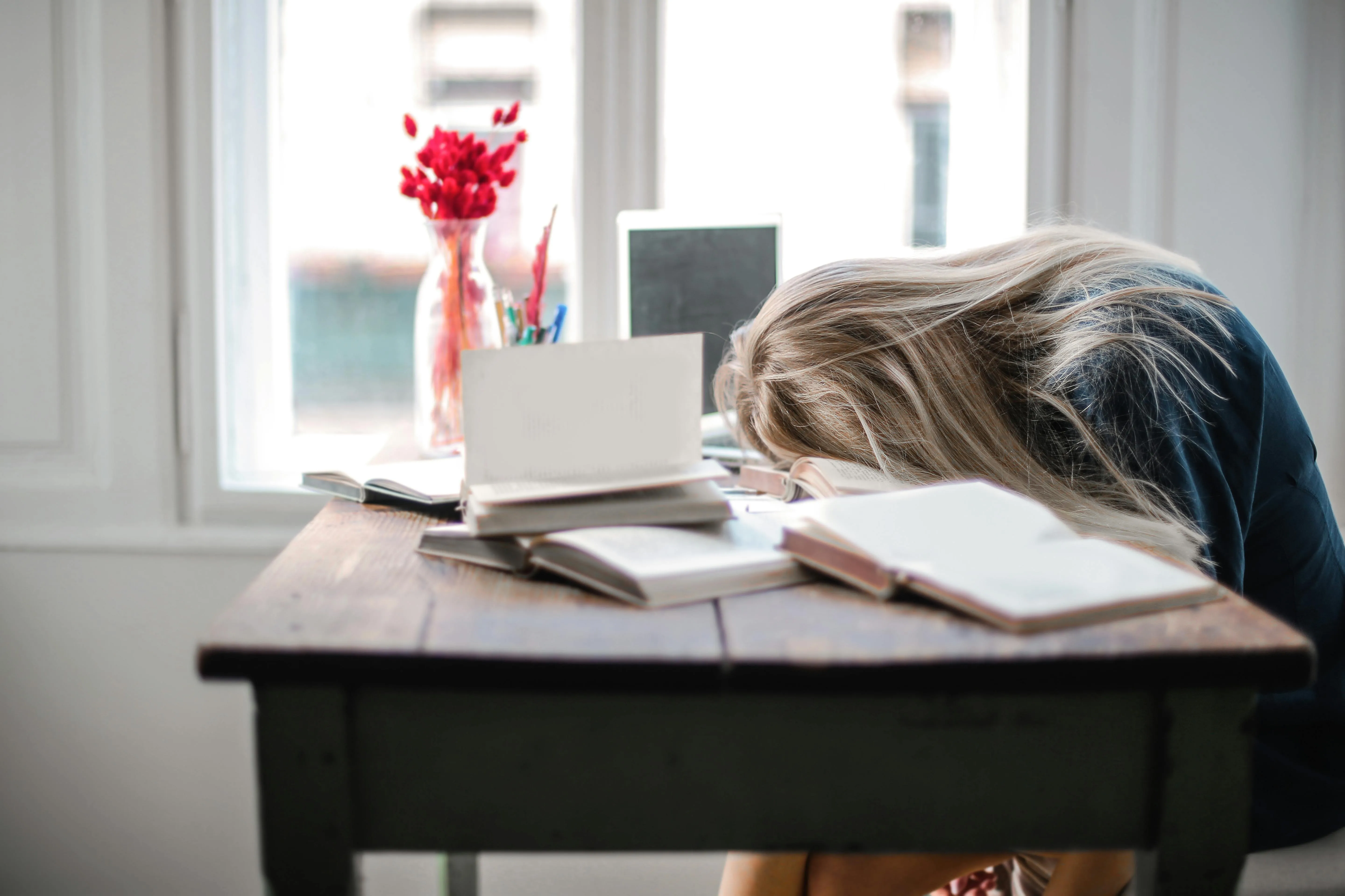 Woman with her head on the table, who is overwhelmed. The table is covered with various books and a laptop.